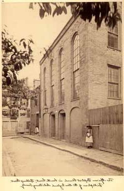 <p>Sepia photograph of a street and the side of a tall, brick building with four arched windows and doors and a large iron lamp. To the left of the building, a Black woman with a shawl, 帽子。, 围裙, and long-sleeved dress stands against a wall with her hands clasped together in front of her.</p>